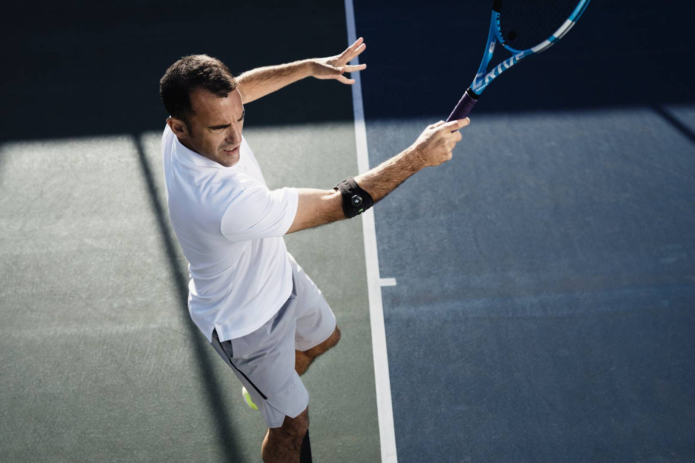 Man playing tennis. He is wearing a white shirt and grey pants. He is also wearing a sports elbow strap, a good support for preventing tennis elbow