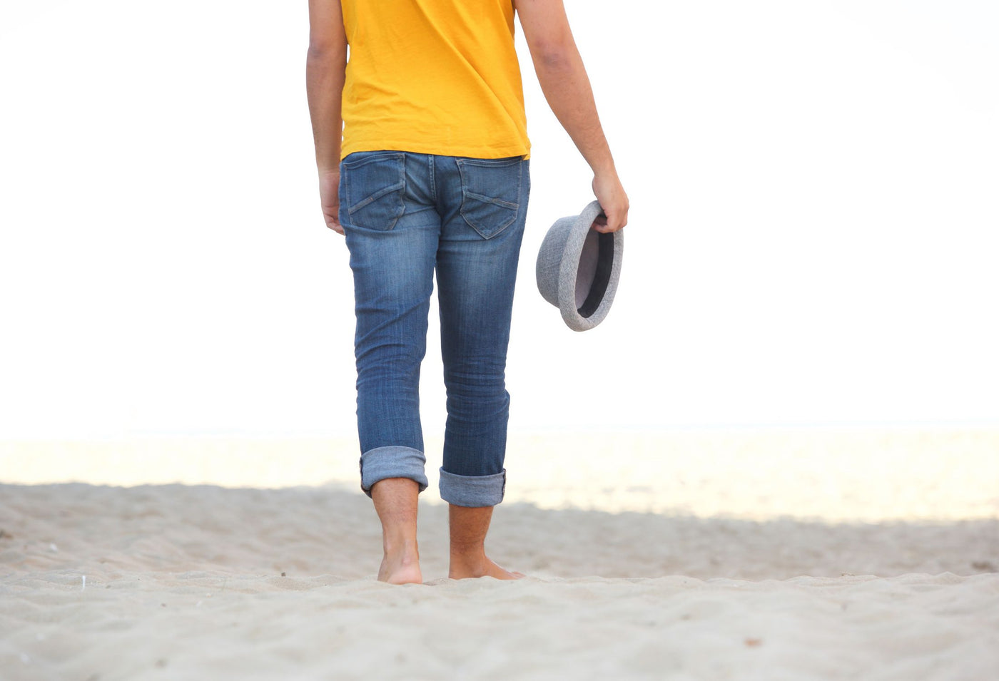 Person walking barefoot on the beach to improve their flat feet. They're wearing a yellow top and blue jeans. 