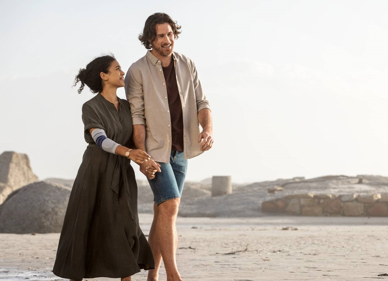 Man and woman walking on the beach, holding hands and talking. The woman is wearing Bauerfeind's EpiTrain elbow brace