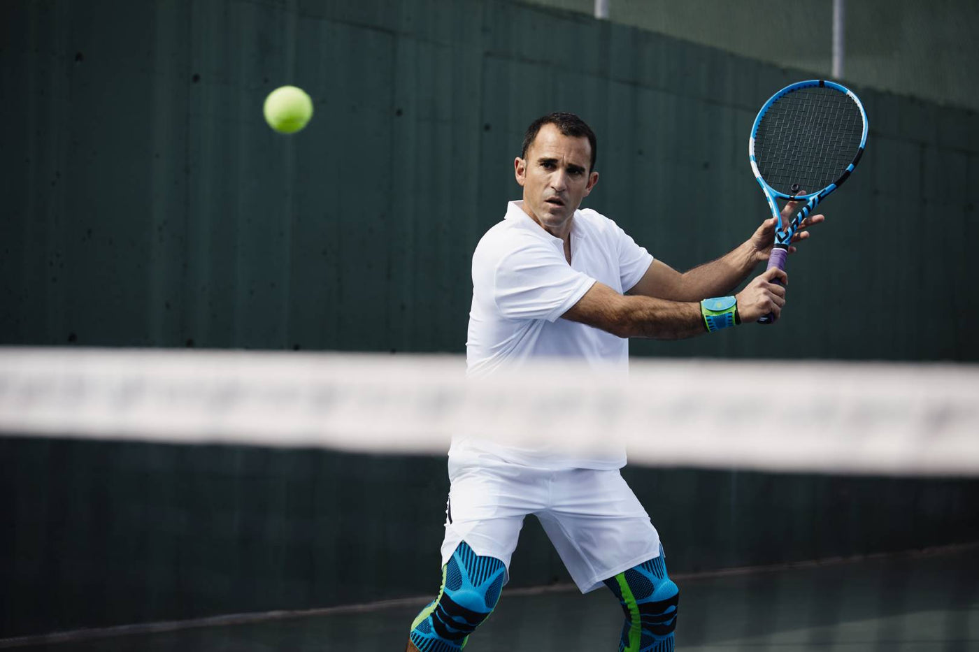Man playing tennis. The tennis net is blurry in the foreground, the man is in focus in the background. He is wearing Bauerfeind Sports Knee Supports and Sports Wrist Strap to avoid tennis injures there.