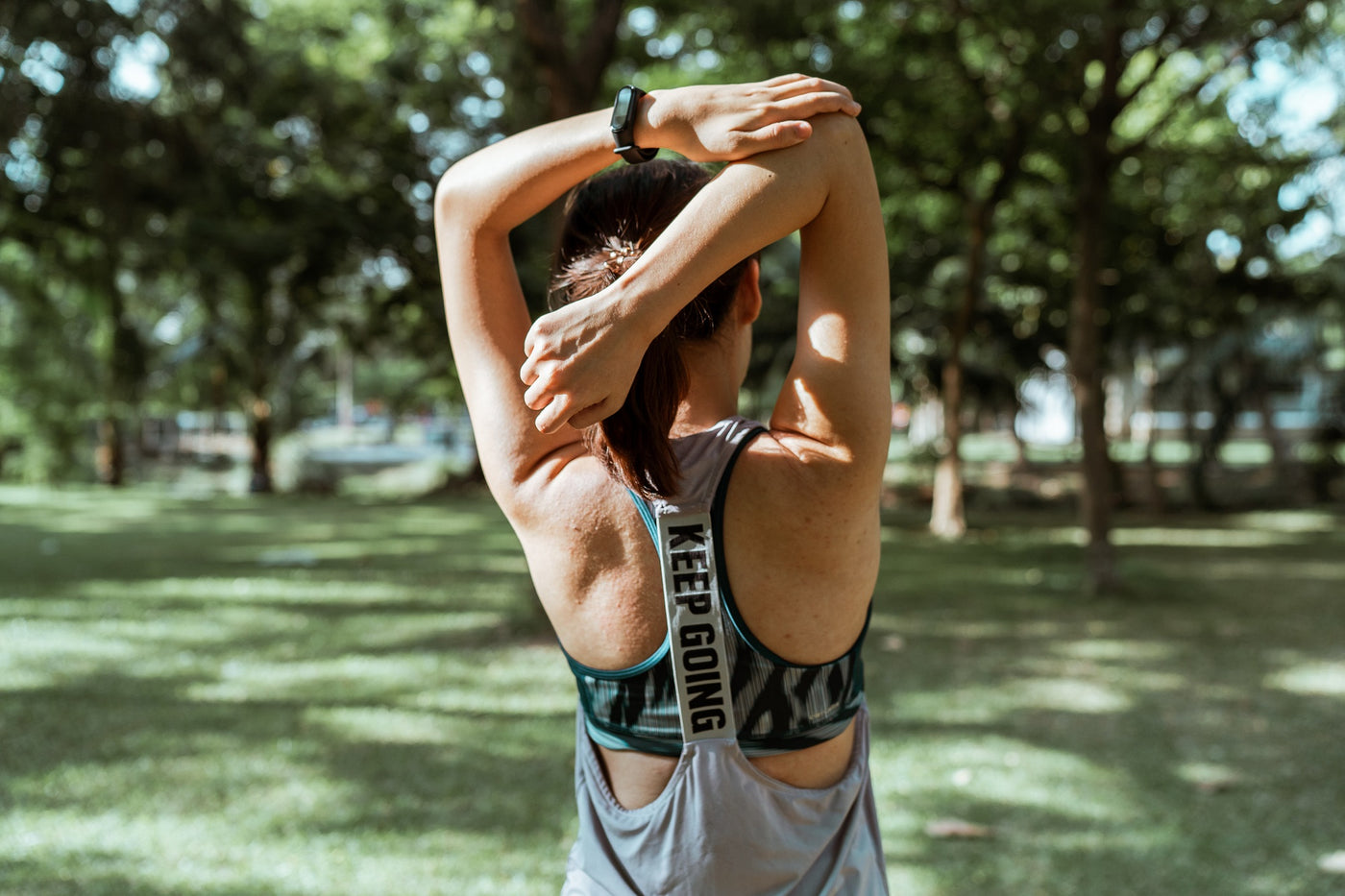 Woman in the park wearing athletic gear and stretching her triceps. The shot focuses on her back and shoulders.