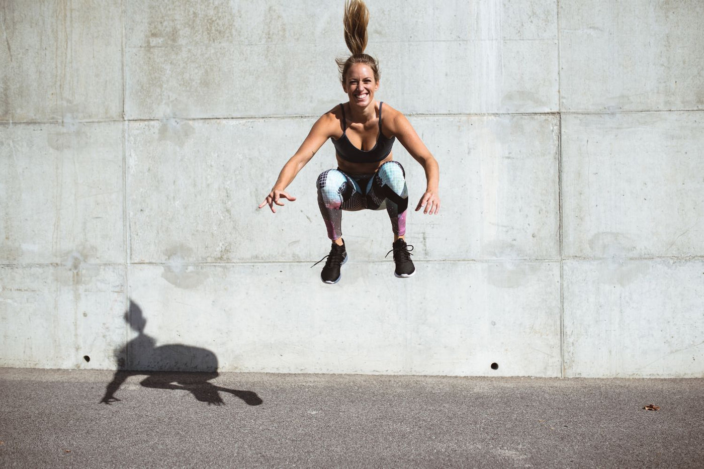Woman doing jump squats in front of a stone wall. She is high off the ground and grinning.