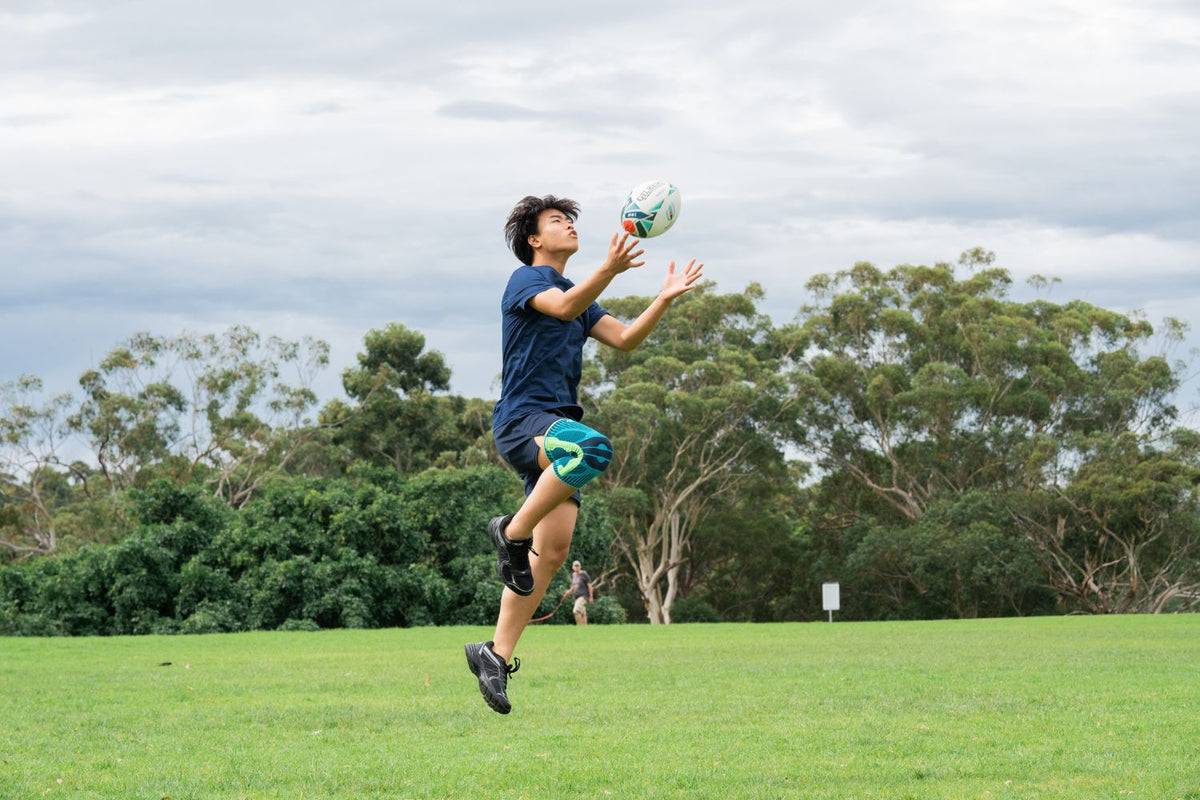 Young man playing ruby at the park. He is jumping up and catching a rugby ball while wearing Bauerfeind's Sports Knee Support
