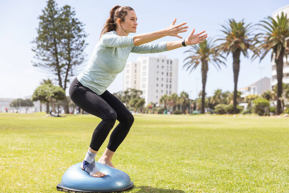 Woman in the park squatting on a balance ball while wearing Bauerfeind's MalleoTrain S ankle brace. Both are great at helping manage ankle tendonitis symptoms 