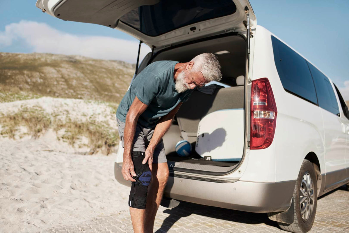 man at the beach standing by the trunk of his car. He is pulling on a knee brace to help manage knee pain 