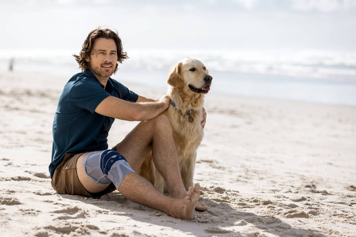 Man sitting with his dog on the beach. He is wearing Bauerfeind's GenuTrain Knee Brace, a good fit for supporting the knee and minimising knee clicking