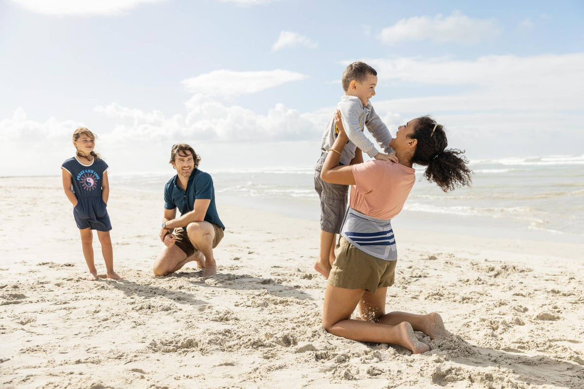 Woman at the beach with her family. She's kneeling and lifting her son up in the air while wearing Bauerfeind's LumboTrain back brace to support her lumbar 