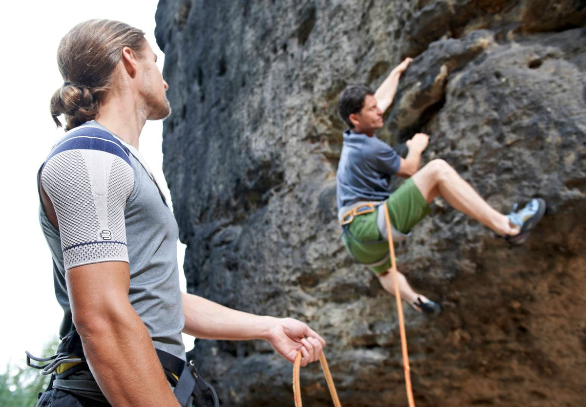 Man holding his freind's rope as the friend climbs up a rock face. The man is wearing Bauerfeind's OmoTrain Shoulder Brace to manage shoulder pain