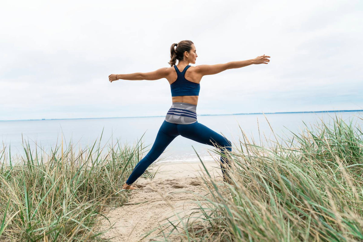 Woman doing yoga on the beach. She is standing in the Warrior pose. She's wearing Bauerfeind LumboTrain Back Brace, one of the best back braces for lower back pain