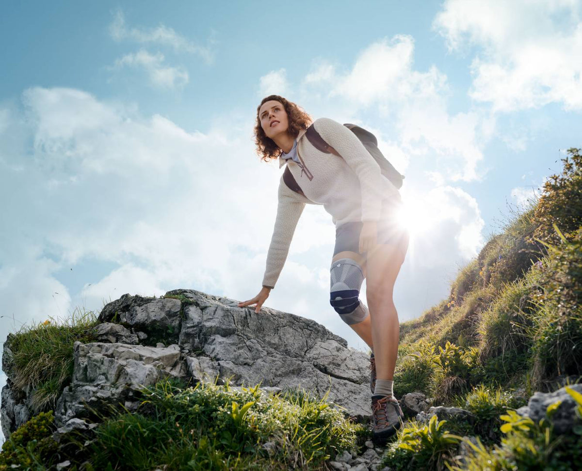 Woman out hiking on a rocky mountain slope. In the shot, the sun is behind her and she faces the camera. She's wearing a GenuTrain Knee Brace, the perfect companion for joint pain relief, injury recovery, and protection.