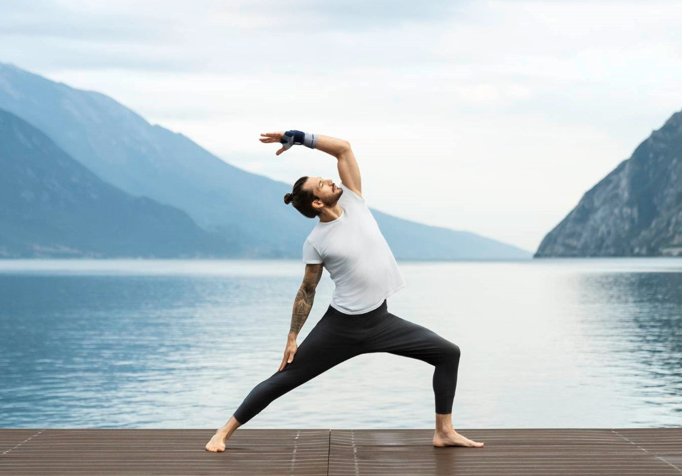 Man with a sprained wrist doing yoga in Bauerfeind's ManuTrain wrist brace. He's exercising on a wooden deck with a lake and mountains in the background