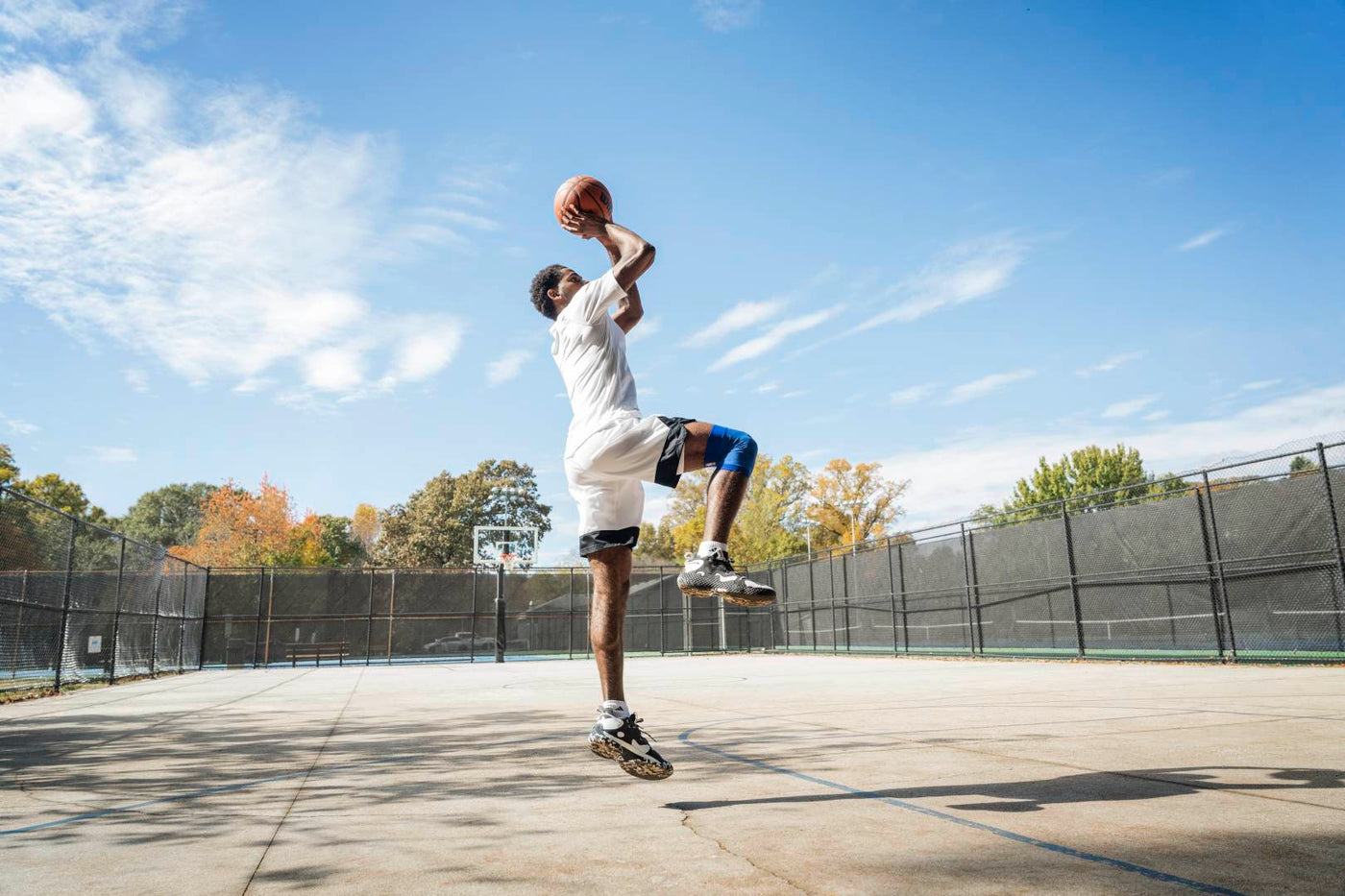 Man in the middle of making a shot on an outdoor basketball court. He is wearing Bauerfeind's NBA Compression Knee Support and basketball shoes, essential basketball training gear