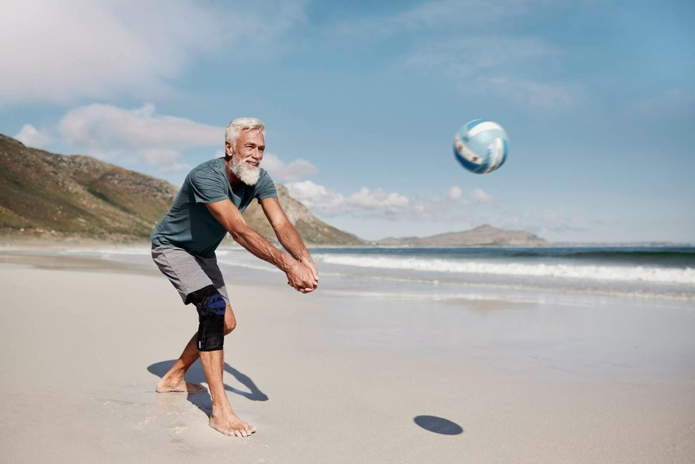 Older man playing volleyball at the beach on a sunny day. He is wearing Bauerfeind's GenuTrain OA osteoarthritis knee brace