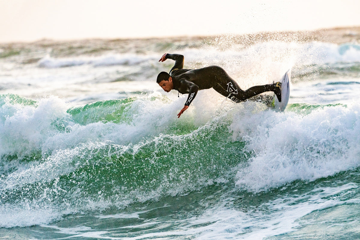 Man in a wetsuit surfing at sunrise. His body is almost parallel to the water, looking as though he is about to lose balance. 