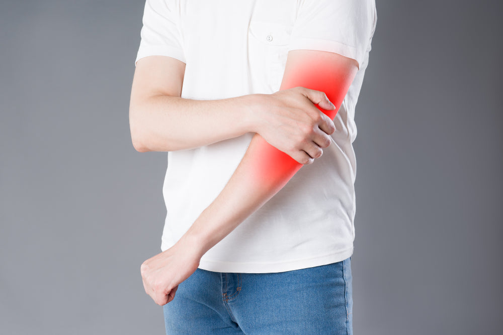 Elbow Pain: Why can’t I straighten my arm? Image of man in white shirt and jeans, he is holding his left elbow and it is highlighted red to indicate pain