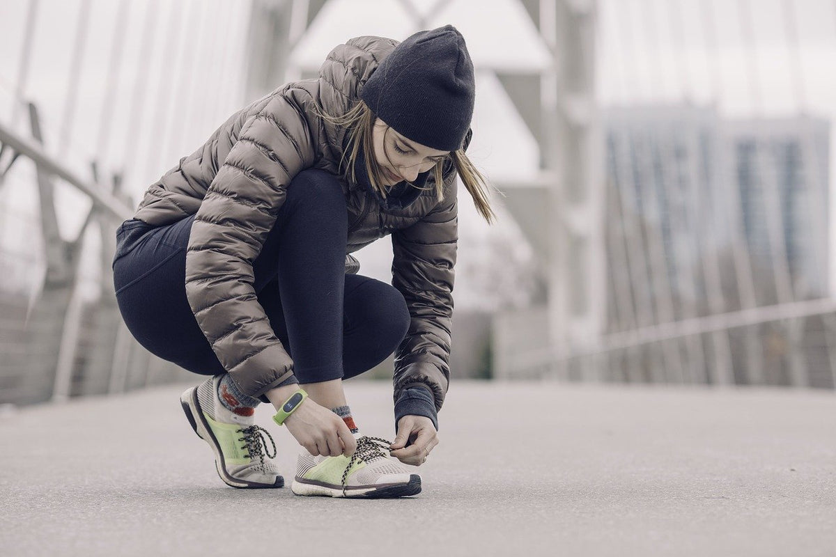 Woman in exercise outfit, warm jacket and beanie. She has stopped to tie her shoelace. The image is related to exercise and stiff joints in cold weather